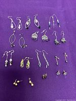 Eleven pairs of assorted earrings arranged on purple fabric, showing different drop and stud styles with various beads and stones.