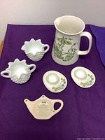 Photo of cream-colored milk pitcher (7 inches high) with green botanical and bird design made in Norway, two white egg cups with green floral motifs, and a small teapot-shaped decorative item with printed text, placed on purple fabric.