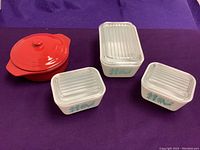 Three white vintage Pyrex bowls with clear plastic lids and red ceramic covered dish with lid on purple background.