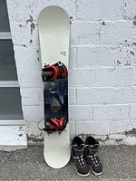 Full snowboard setup leaning against white wall, showing snowboard, bindings attached, boots at base.