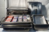 Multiple stainless steel catering trays stacked with food warmer trays with lids; includes boxes of cooking fuel canisters on top.