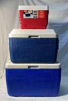 Three coolers stacked: two blue Coleman coolers with white lids at bottom and middle, and a small red Igloo cooler with white lid on top