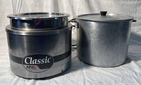 APW Classic crockpot and large aluminum pot with lid side by side on a white backdrop.