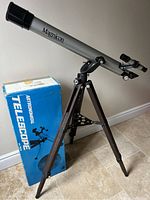 Magnicon Stargazer 234 telescope assembled on wooden tripod with original box standing behind it on tile floor against wall.
