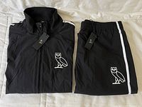 Full view of folded black track jacket and matching track pants with white owl logos and tags attached