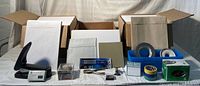 Photo of the full lot showing card stock, envelopes, multiple rolls of tape, a stapler with staples, rubber bands, paper clips, and other small office supplies on a white surface with open boxes in the background.