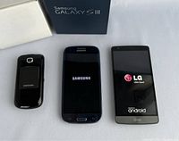 Three cell phones side by side: Samsung Galaxy S III, LG Android smartphone, and Samsung flip phone with their systems booting or resting screens.