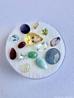 Front and top view of various cut gemstones placed on round white foam inside a plastic container showing colors and shapes