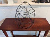 Black metal polygonal geometric cage for pendant light placed on wooden table, showing overall shape and design.