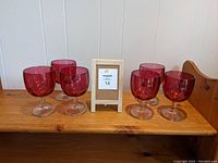 Six red-tinted large wine goblets with clear stems and bases displayed on a wooden surface.