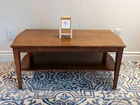 Front view of rectangular solid wood coffee table on patterned rug, showing built-in lower shelf and tapered legs