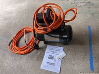 Air compressor unit with attached orange air hose and power cord, shown with instruction manual on concrete floor.