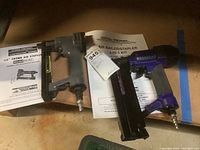 Two air nailers displayed on a brown surface with manuals, showing the Central Pneumatic brand and model numbers 6365 and 4916.