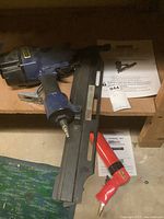 Photo showing the blue framing nailer and the red pneumatic caulking gun along with manuals placed on a wooden shelf.