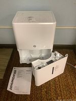 White GE dehumidifier unit with front water tank detached, placed on carpet, along with user manual.