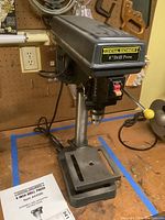 Full view of Central Machinery 8-inch drill press showing base, worktable, motor housing, and power cord with manual visible on the workbench.