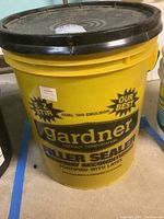 Yellow 5 gallon bucket labeled Gardner drive way filler sealer with black lid on concrete floor.