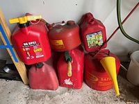 Photo showing six gas cans together, four plastic red cans with one missing lid, two red metal cans including one with yellow SAFE label and a yellow funnel attached.