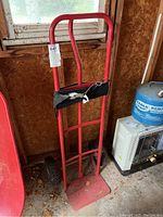 Hand truck standing upright against a wooden wall, showing red metal frame and base plate with attached black strap.