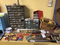 Wide view of workbench with multiple small parts organizers on the table containing screws, bolts, and other hardware. Hand tools, packs of staples, and miscellaneous items arranged on the work surface.