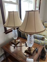 Two matching table lamps with clear cut glass bodies and square gold-toned metal bases, each topped with beige fabric bell shades. Lamps sitting on wooden table near window.