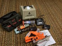 Full view of the power tools lot showing circular saw, belt sander, batteries, charger, bags and cases on carpet.