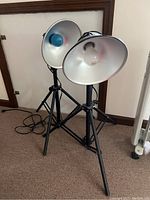 Two black tripod lights with large metal reflector shades, one with a blue bulb, standing side by side indoors on carpet.