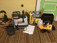 Overview of Wagner FineCoat and Wide Shot spray painting units with cords, hoses, packaging, and manuals on carpeted floor in front of green door