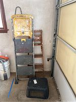 Yellow Cosco metal step ladder, wooden A-frame ladder, and black plastic step stool together showing overall lot contents
