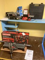 Craftsman belt sander, disk sander, hard case on blue table, and boxed miter box below