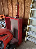 Side view of the red steel tow behind cart adjacent to a Kubota lawn tractor in a wooden shed.