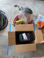 View showing all items: welding helmet inside box, clear face shield with headband, red hose with brass torch end, yellow gas canister, and some background items.