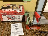 Tile saw on carpet next to its original box and instruction manual.