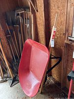 Red 4 cu ft wheelbarrow with wooden handles and black metal support frame in a shed, alongside other garden tools.