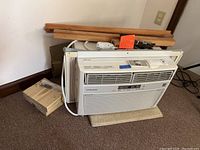 Frigidaire window air conditioner unit resting on carpet with some wooden mounting accessories behind it.