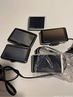Five Garmin GPS units displayed on white surface. Various screen sizes and bezel colors visible, two units connected to cables, one wrapped in plastic.