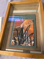 Framed abstract art piece with a glass front inside a light wood frame. The art features black, white, and orange brush strokes.