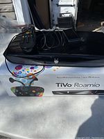 Black TiVo Roamio DVR on colorful box with remote control and power adapter on top.