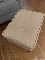 Top view of beige fabric upholstered ottoman with piping detail on edges and wooden legs.
