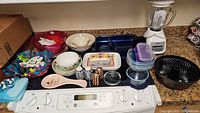 Wide view of counter showing all items in the lot: blue Pyrex baking dishes, Lenox and Royal Worcester serving pieces, black bundt pan, plastic storage containers, Oster blender, and decorative items.