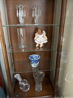 Wooden display cabinet shelf with two crystal candlestick holders with hanging prisms on top shelf, clear glass cylinder vase and wooden vintage doll on middle shelf, blue and white Asian candle warmer and crystal vase on bottom shelf.