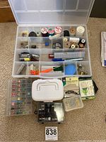Top view of plastic organizer box filled with assorted small sewing supplies including thread spools, bobbins, pins, needles, and tools plus several small plastic containers including a bobbin winder.