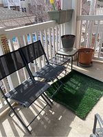 Two black folding chairs with mesh seats and backs positioned beside a small table on a balcony with a green faux grass rug and several plant pots.