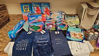 Overview of food storage bags, trash compactor bags, parchment paper, plastic wrap, and three navy blue kitchen towels with printed messages.