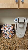 Wide photo showing white Keurig coffee maker next to rotating metal stand with multiple coffee pods on granite countertop.