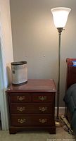 Full view of wooden nightstand dresser with gold floor lamp and decorative trash bin on top, next to bed.