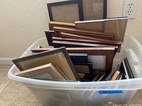 Two plastic storage bins filled with roughly 30 assorted picture frames of various sizes, materials, and colors. Some frames missing glass or plexiglass inserts.