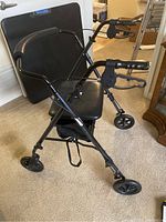 Folded four wheel walker displaying black padded seat, hand brakes, and wheels. Walker folded slightly and standing up.