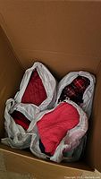 Three white plastic bags placed inside a large cardboard box, containing folded sweaters, one with a red quilted detail and visible red and black plaid fabric in one bag.