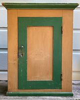 Front view of the antique pine hanging wall cabinet, showing the closed single door with green and natural pine paint, lock and keyhole visible.